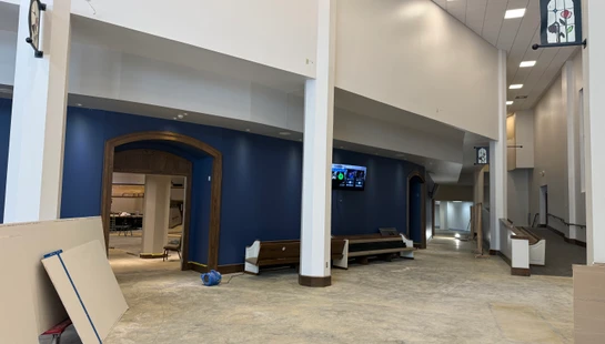 A large church lobby with white pillars and a new blue accent wall is under construction, with dusty concrete floors and drywall sheets leaning against a wall.