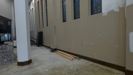 A large hallway in a church with concrete floors and beige walls mid-renovation, with stained glass windows and construction materials on the floor.