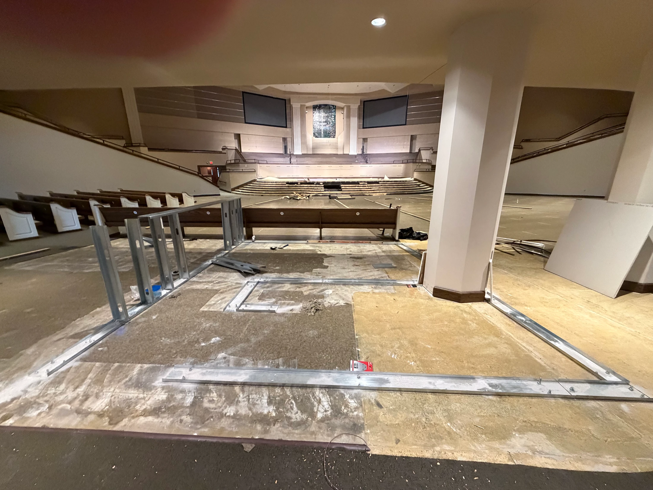 A wide view of a large church auditorium during renovation, with metal framing on the floor in the foreground and pews and a stage in the background.
