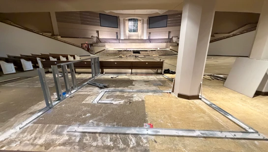 A wide view of a large church auditorium during renovation, with metal framing on the floor in the foreground and pews and a stage in the background.