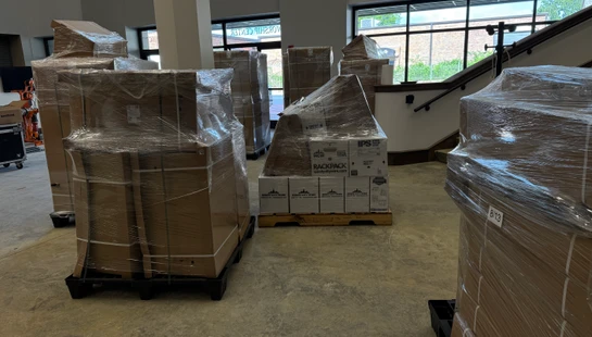 A room with several large pallets of shrink-wrapped cardboard boxes. In the background is a staircase and a window with a 'WORSHIP CENTER' sign visible.