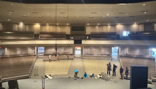 A wide, elevated shot of a large auditorium with pews where workers are on the main floor, appearing to be removing or installing carpeting.