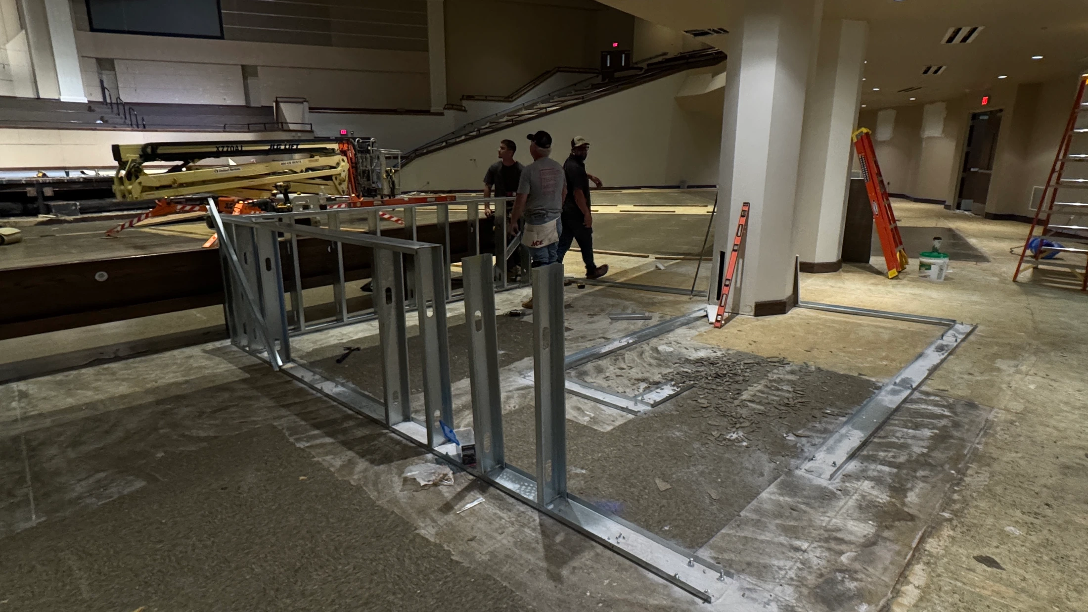 Three construction workers stand near newly installed metal framing for walls in a large, open auditorium undergoing extensive renovation.