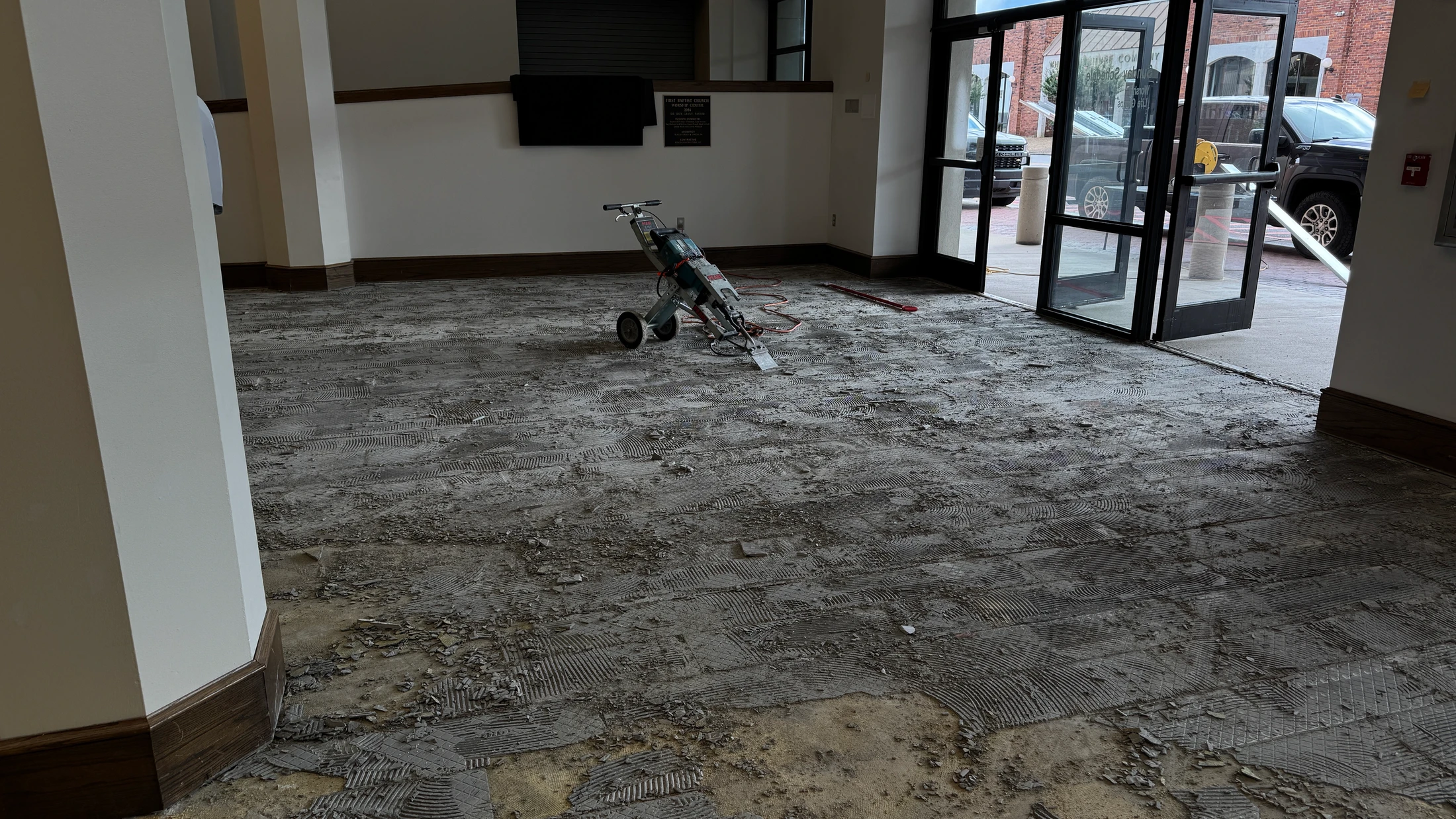 The floor of a building lobby is torn up, showing old adhesive and debris. A floor scraper machine sits in the middle of the room during a renovation.