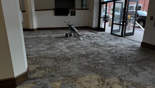 The floor of a building lobby is torn up, showing old adhesive and debris. A floor scraper machine sits in the middle of the room during a renovation.
