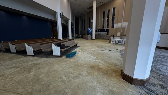 A church sanctuary undergoing renovation. Pews are pushed to one side, and the old carpet has been removed, exposing the bare concrete subfloor.