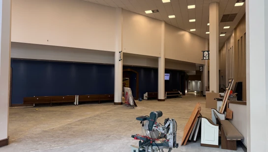 A large, empty hall undergoing renovation, with a miter saw in the foreground on a bare concrete floor and a dark blue accent wall in the background.
