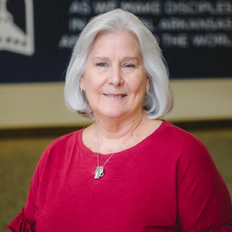 A head and shoulders portrait of a smiling woman with short silver hair and blue eyes. She is wearing a red shirt and a simple silver necklace.
