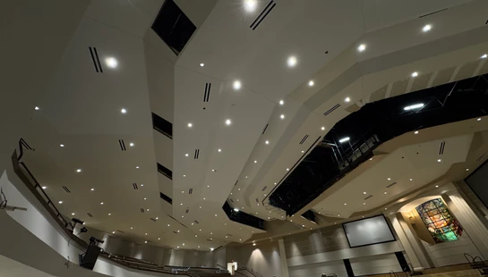 Wide-angle view looking up at a complex, modern ceiling in a large auditorium during construction, with many recessed lights, vents, and a stained glass window.