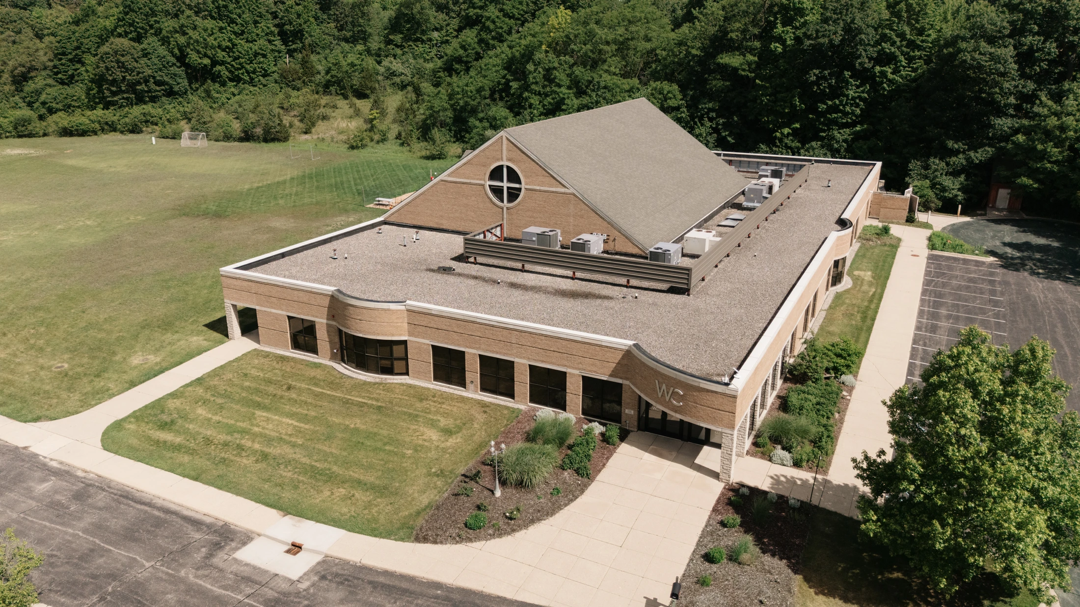 An aerial view of a modern, tan brick building with a partial pitched roof, surrounded by green lawns, a parking lot, a soccer field, and a dense forest.