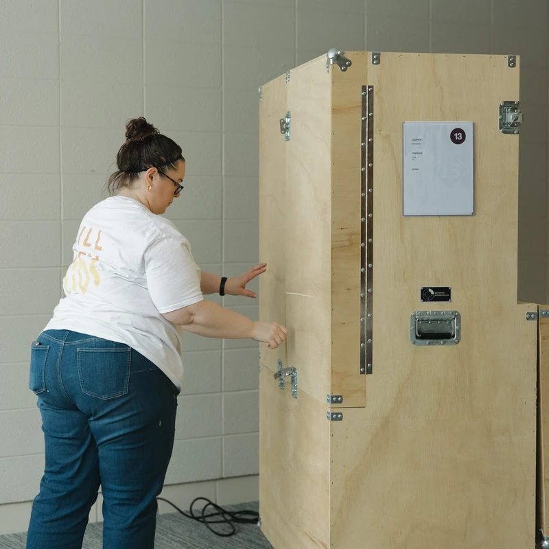 A woman wearing glasses and jeans stands indoors next to a tall wooden shipping crate on wheels, reaching out to touch its side near the latches.