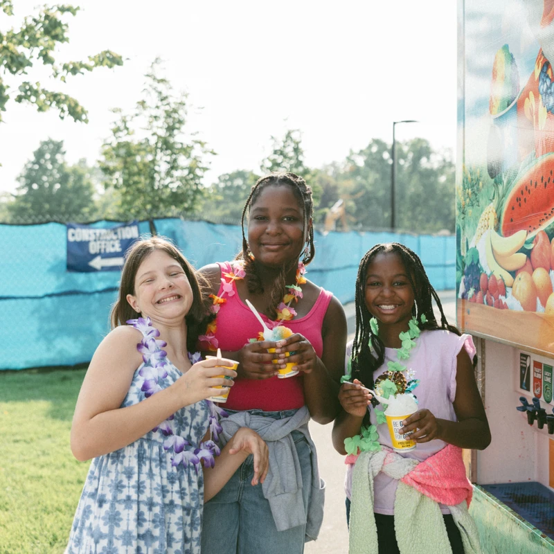 Three diverse young friends wearing colorful flower leis smile while holding cups of shaved ice outdoors on a sunny day by a beverage dispenser.