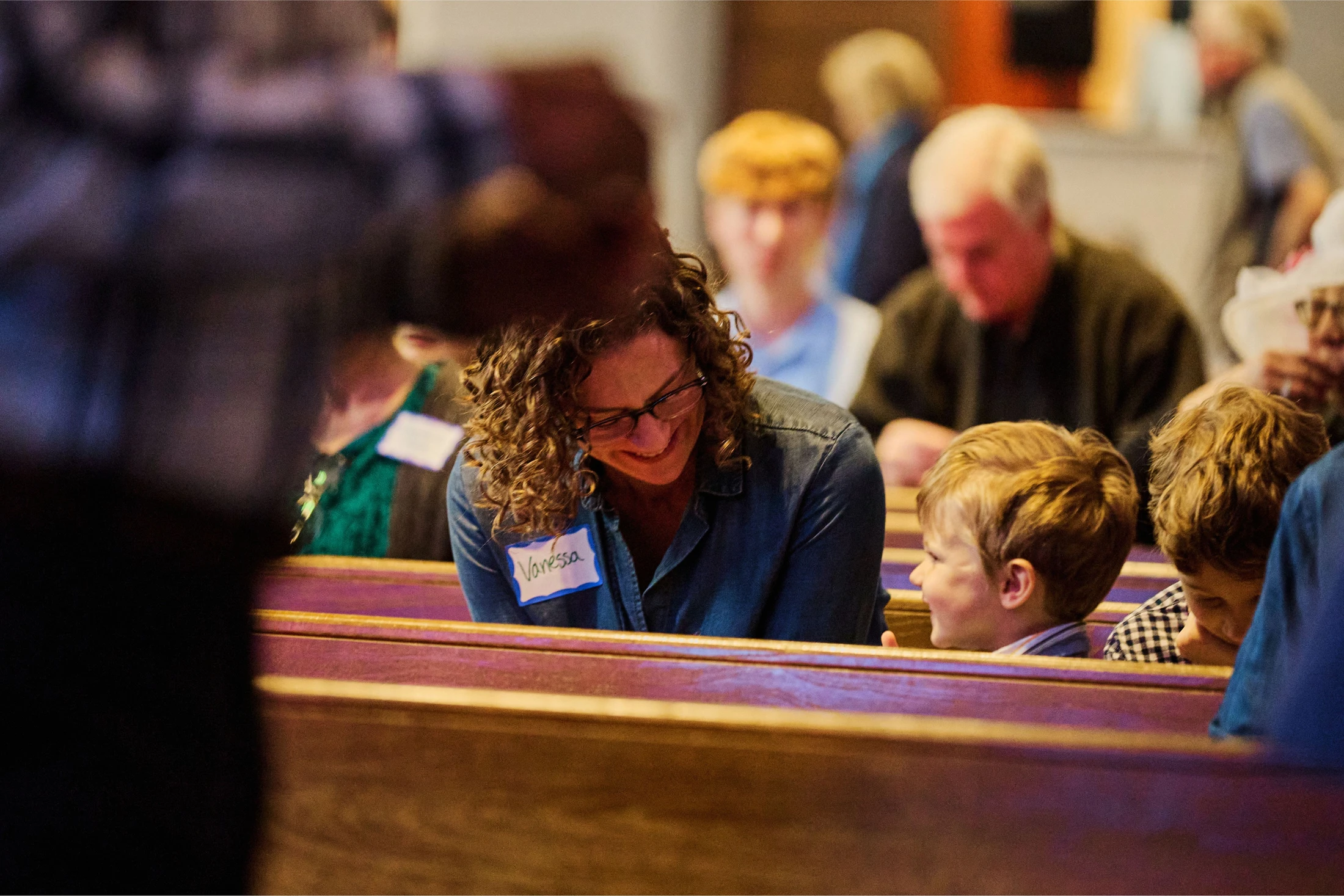 A woman with curly hair and a name tag smiles down at a young boy next to her as they sit together on a wooden pew in a crowded room.
