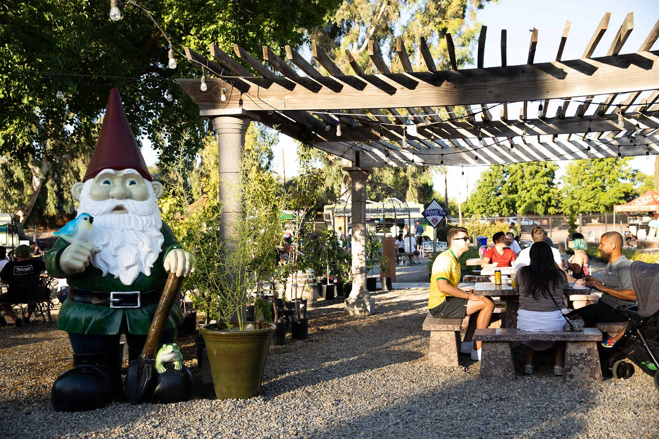 A large garden gnome statue stands on a gravel path next to people sitting at picnic tables under a wooden pergola with string lights.