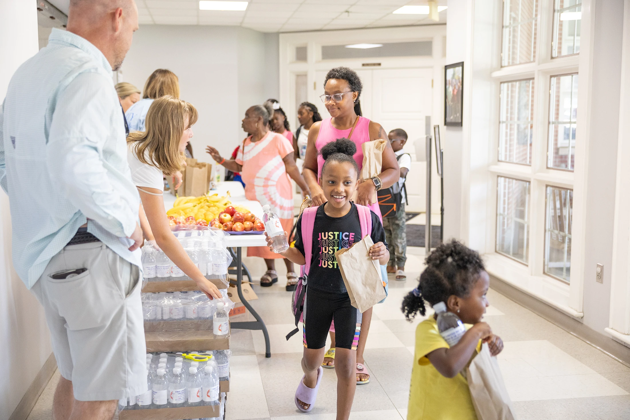 A young Black girl with a pink backpack smiles as she gets a water bottle and a paper bag at an event where volunteers hand out refreshments to families.