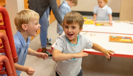 Two young boys laugh and play together in a classroom, while another boy, a girl, and a teacher are in the background near a table with puzzles.