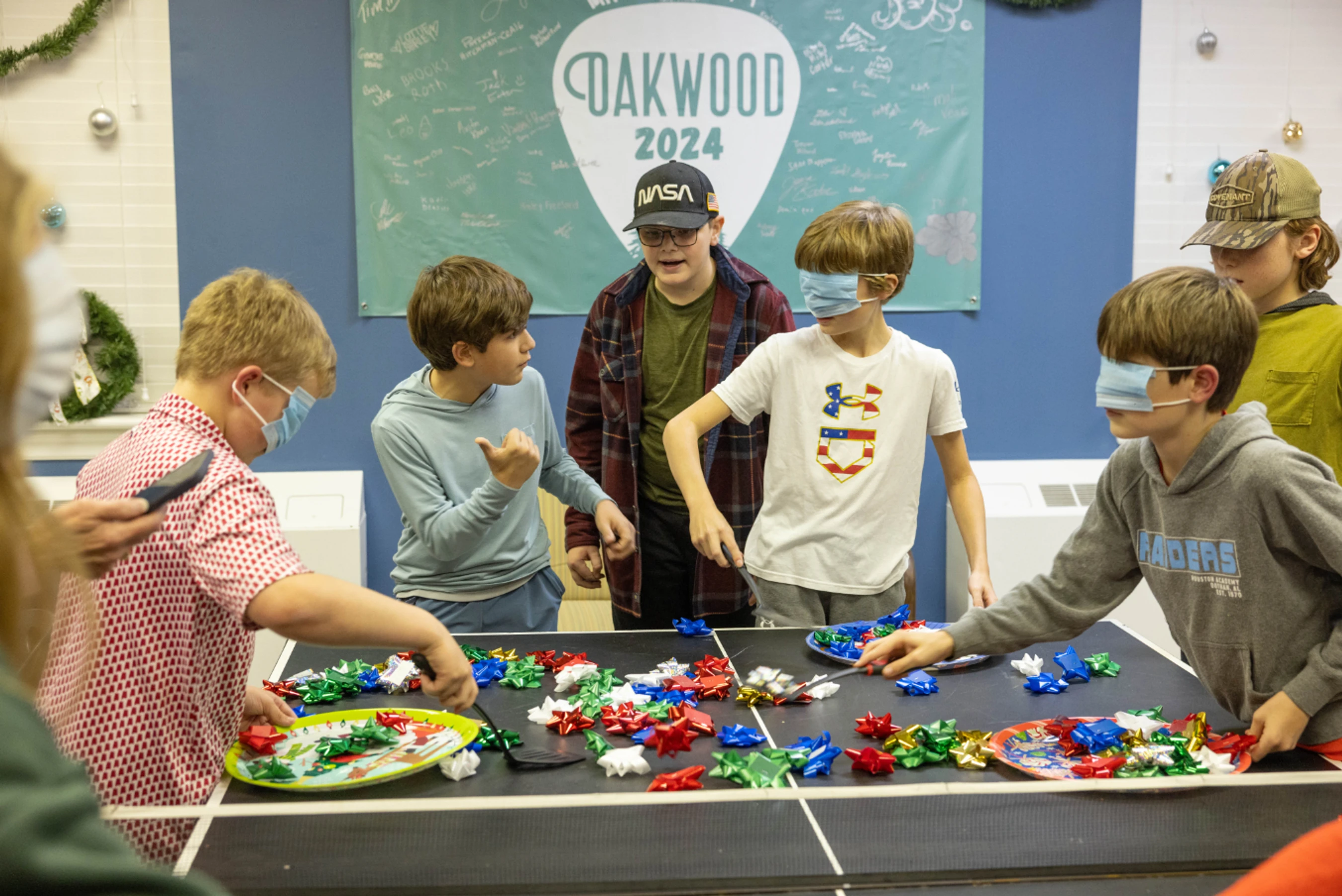 A group of young boys play a game at a holiday party. Two boys wear face masks as blindfolds while trying to scoop colorful bows onto plates with spatulas.