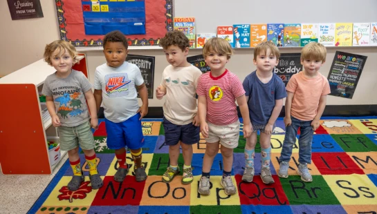 Six diverse young boys stand in a line for a group photo in their classroom. They are on a colorful alphabet rug with children's books in the background.