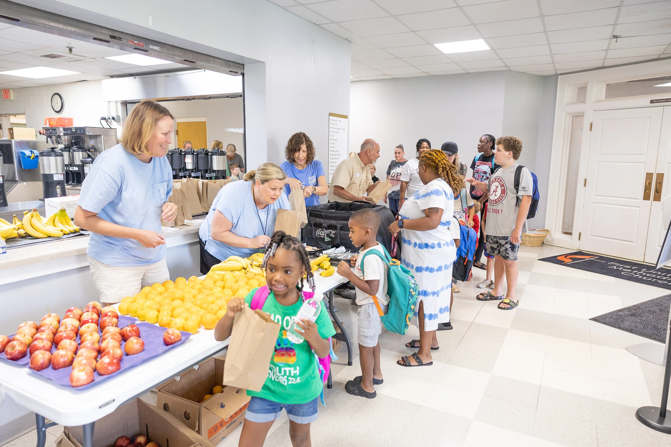 Volunteers hand out bags of fresh fruit, like apples and oranges, to children and adults waiting in line inside a community hall.
