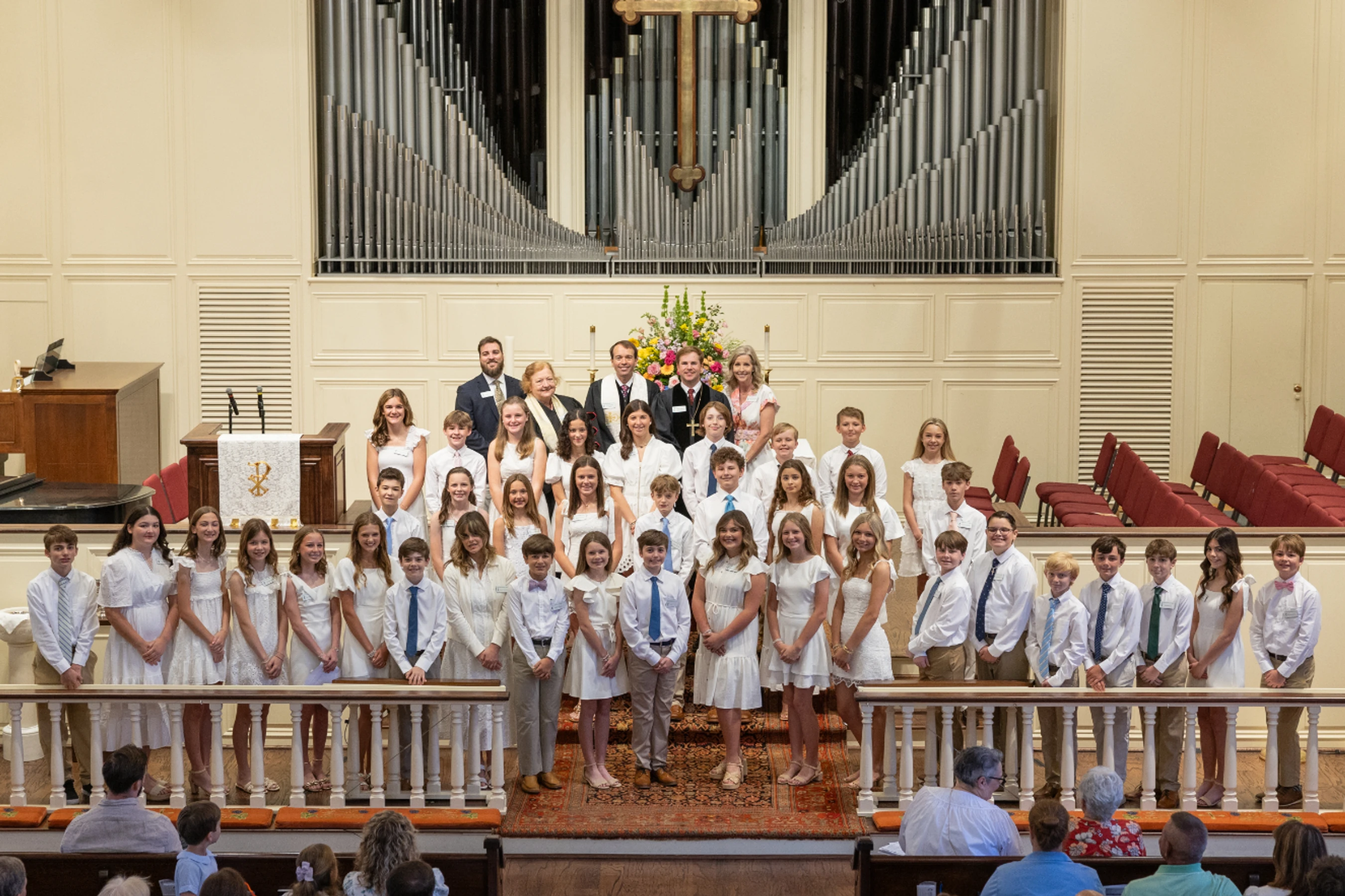 A large group of children and adults pose for a formal photo at the altar of a church, in front of a large pipe organ and a cross.