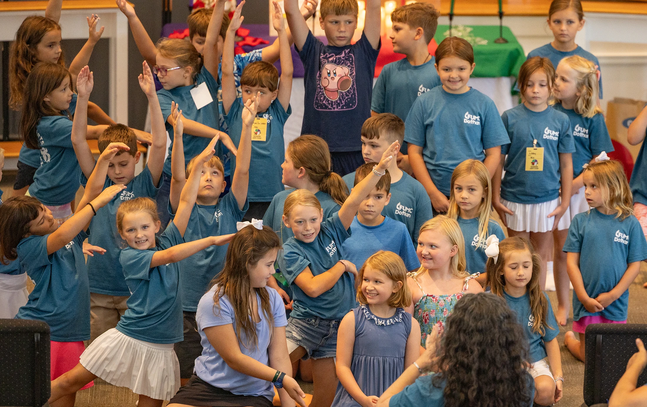 A large group of elementary-aged children in matching blue t-shirts participate in an activity, with many raising their hands in the air enthusiastically.