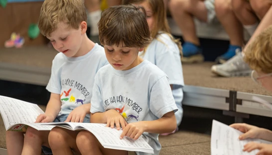 Two young boys in matching light blue shirts sit side-by-side, intently reading from a book of sheet music on their laps. One boy points to the notes.