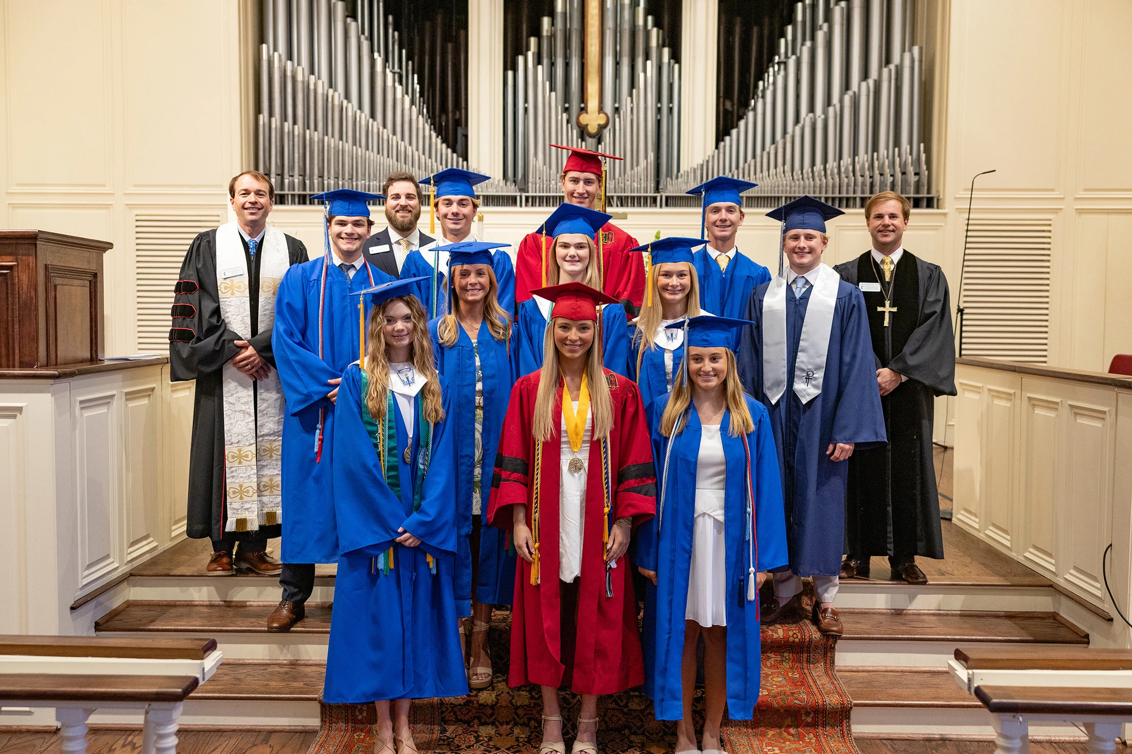A group of graduates in blue and red caps and gowns pose for a photo with faculty on the steps of a church altar, with large organ pipes in the background.