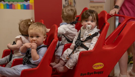 Four babies are buckled into a large red multi-seat stroller inside a classroom. One baby has a pacifier and another looks at the camera.