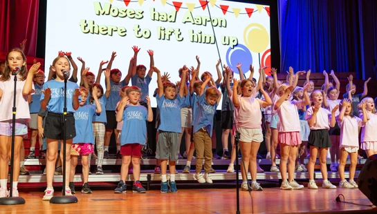 A large group of young children sing on a wooden stage with their hands raised. A screen behind them displays song lyrics for a church performance.