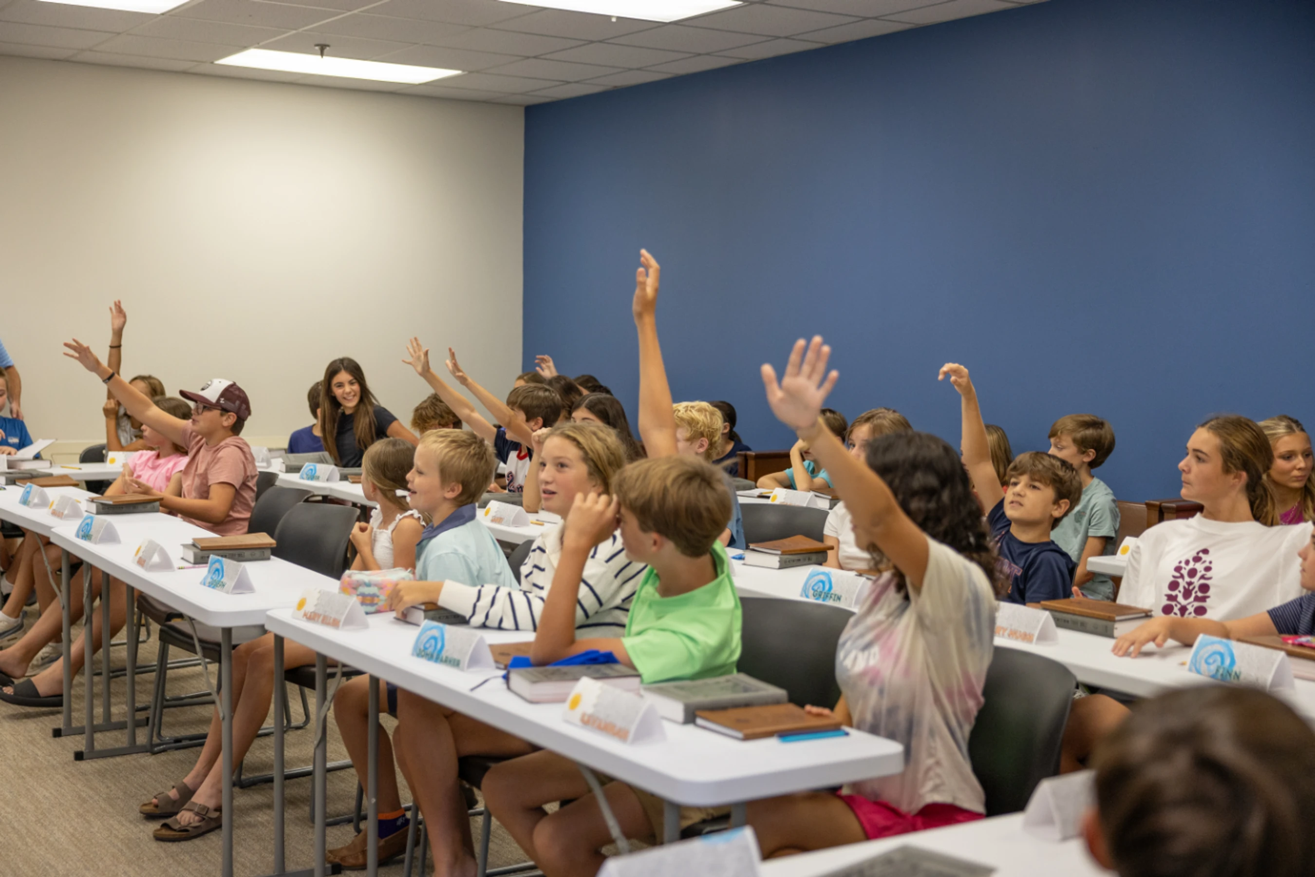 A classroom full of young students sitting at long white tables with their hands raised, eager to participate in a lesson.