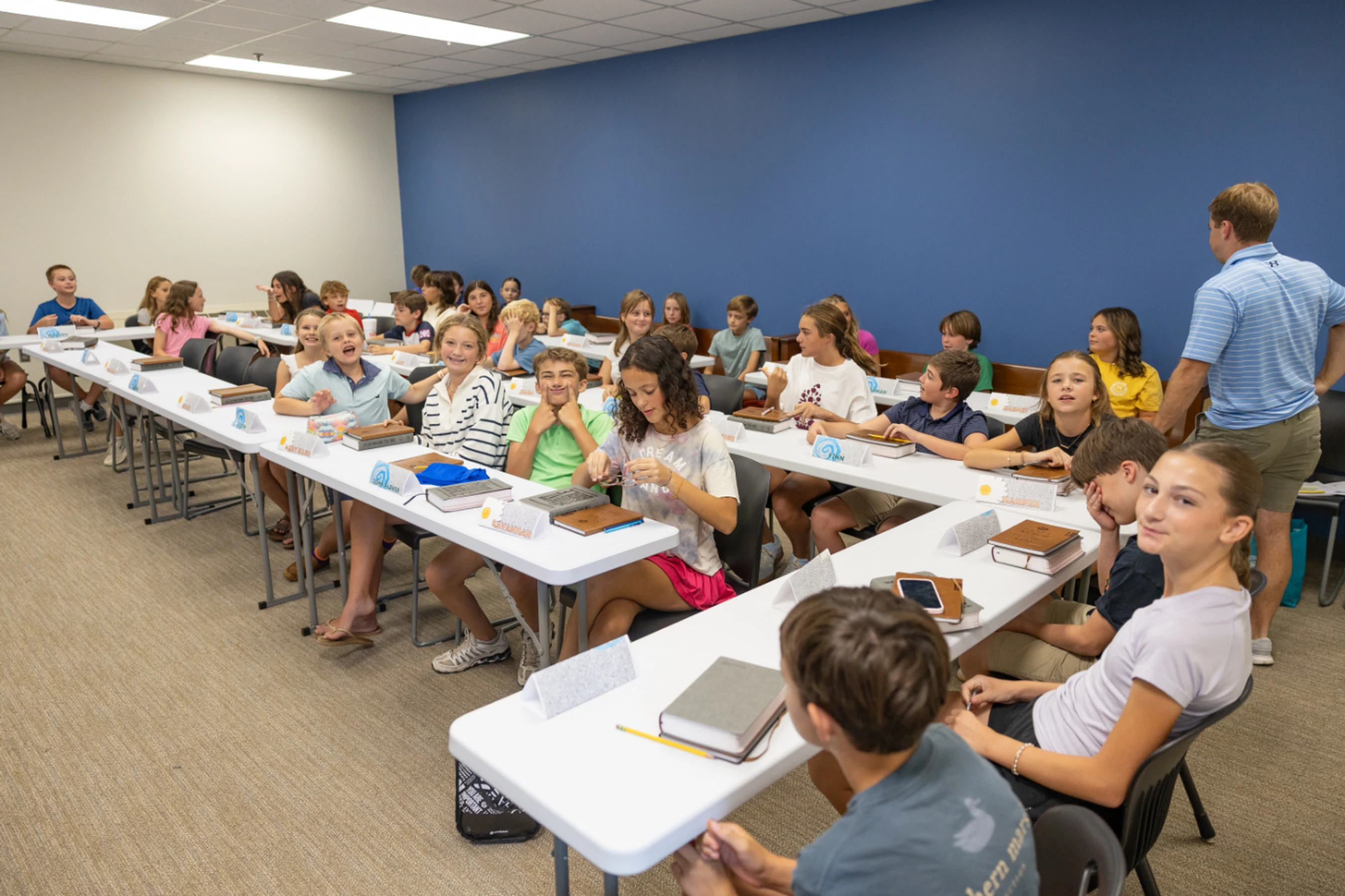 A large group of middle school-aged students sits at long white tables in a classroom with a blue wall, interacting and smiling during a lesson.