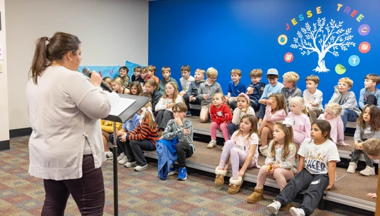 A woman stands at a podium with a microphone, speaking to a large group of elementary school children sitting on risers in front of a blue wall.