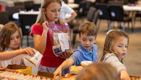 A group of four young children stand around a table, concentrating as they pack snacks and food items from cardboard boxes into clear plastic donation bags.