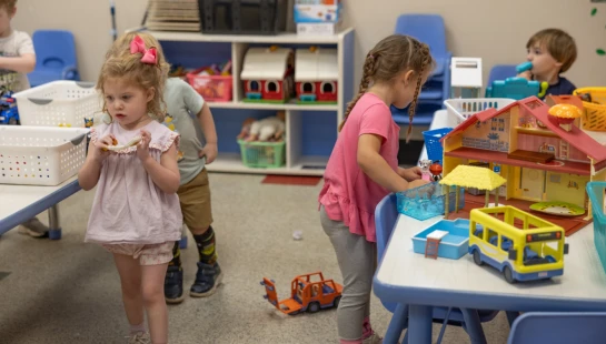 A group of young children in a classroom setting, with one girl playing with a dollhouse and another holding a toy, surrounded by shelves of toys.
