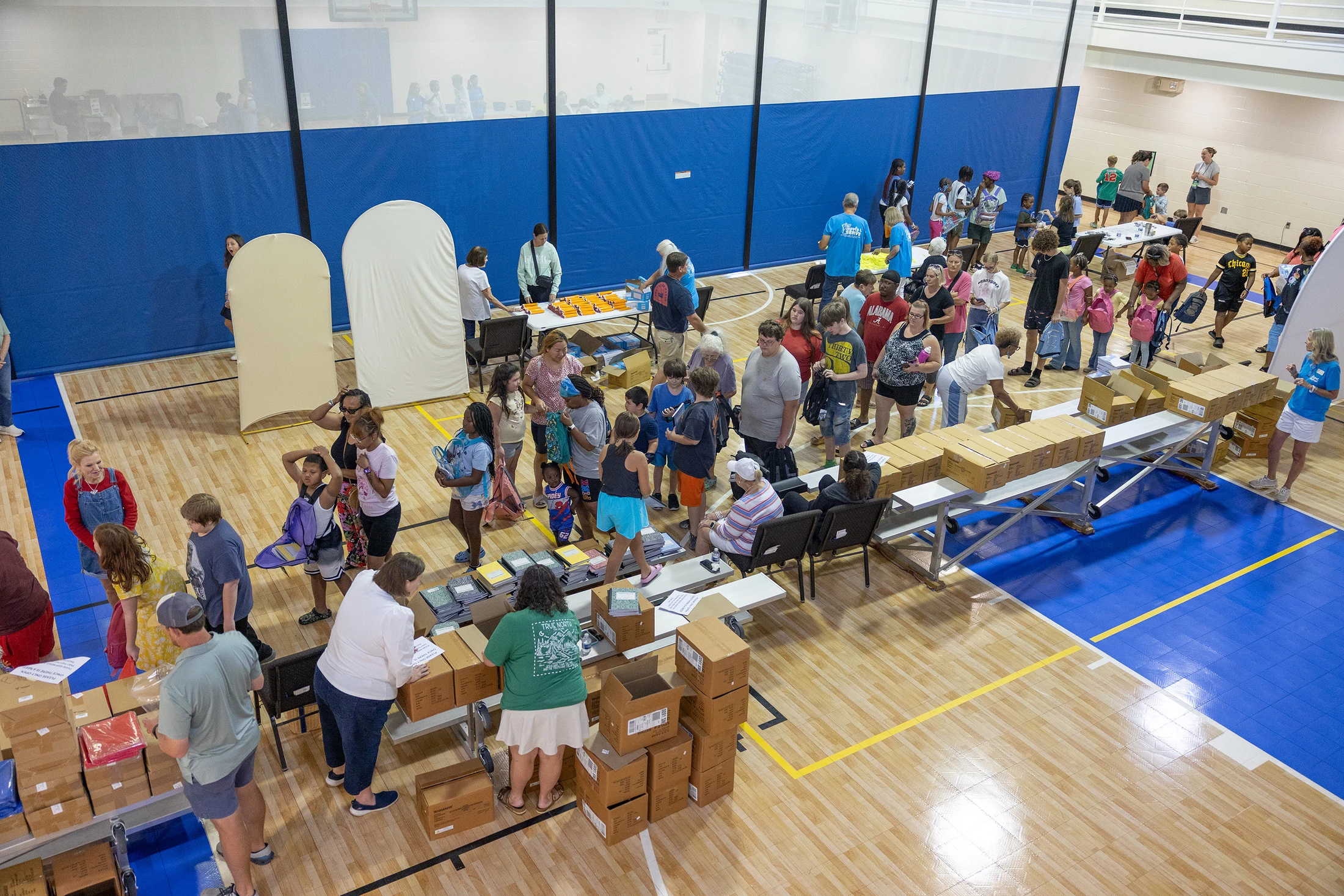 A high-angle shot of a busy gymnasium where families and children line up at tables to receive school supplies distributed by volunteers.