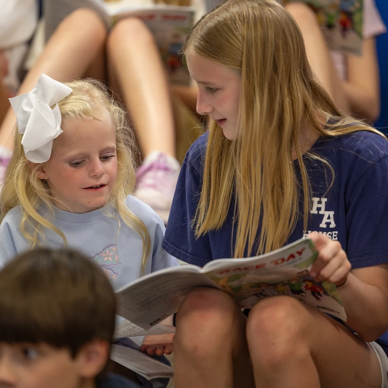 An older girl with blonde hair reads from a picture book to a younger girl with a large white bow in her hair. Both are sitting on the floor.