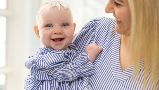 A happy baby girl with a white bow smiles at the camera while being held by her mother. They are both wearing matching blue and white striped outfits.