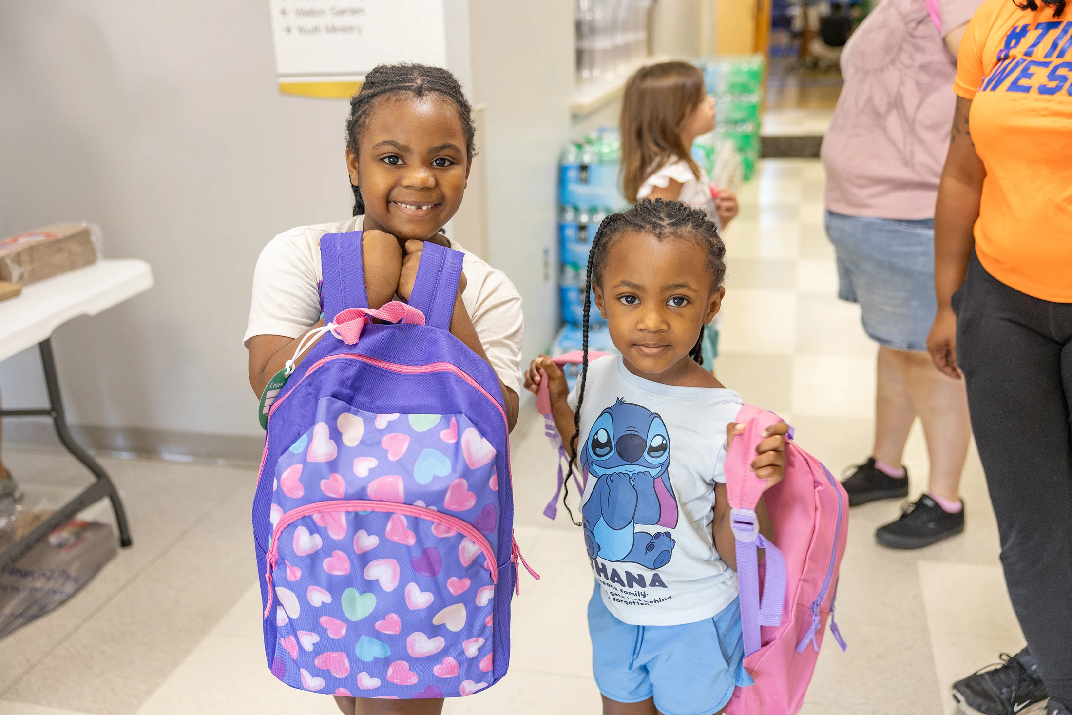 Two young Black girls stand side-by-side holding new backpacks. The older girl smiles while holding a purple backpack, and the younger girl holds a pink one.