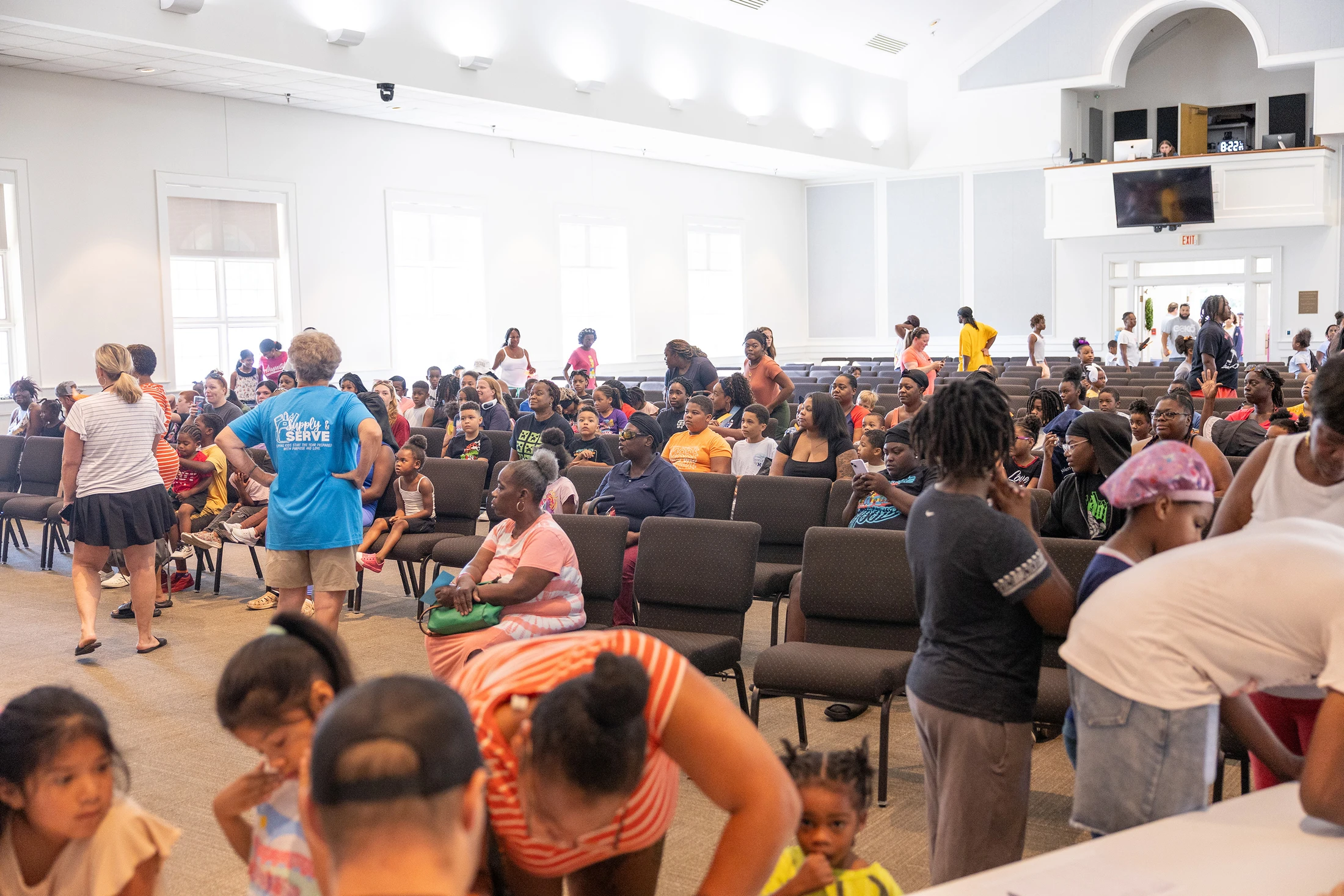 A large, diverse group of families and children gather inside a modern hall for a 'Supply & Serve' community event, sitting in rows of chairs.