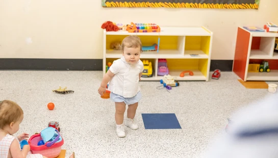 A toddler in a white shirt and blue shorts stands in a playroom, looking at the camera. Another child plays on the floor and toys are scattered around.
