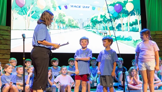 A woman with a clipboard talks to three children standing on stage in front of other seated kids. A screen behind them shows a cartoon 'Race Day' scene.