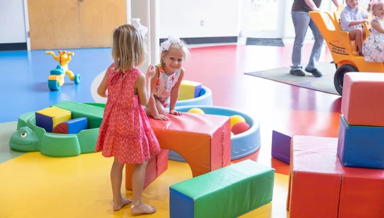 Young children play in a bright, colorful room. Two girls are near large soft blocks, while an adult pushes other children in a multi-seat wagon.