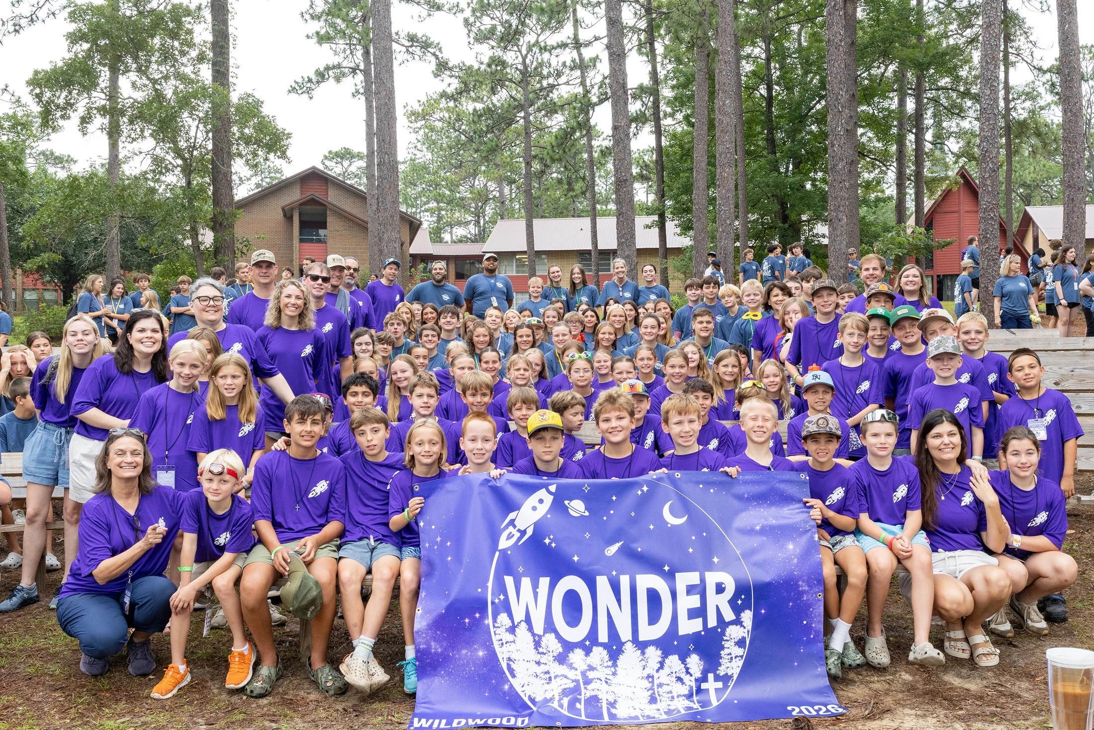 A large group of children and adults in matching purple shirts pose for a photo at a wooded camp, holding a blue banner that reads 'WONDER' and 'WILDWOOD'.