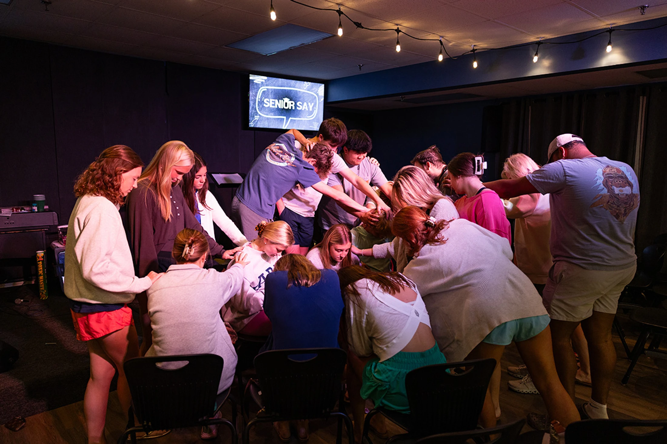 A large group of young people huddle in a circle, praying with hands on each other's shoulders under dim string lights during a 'Senior Say' event.