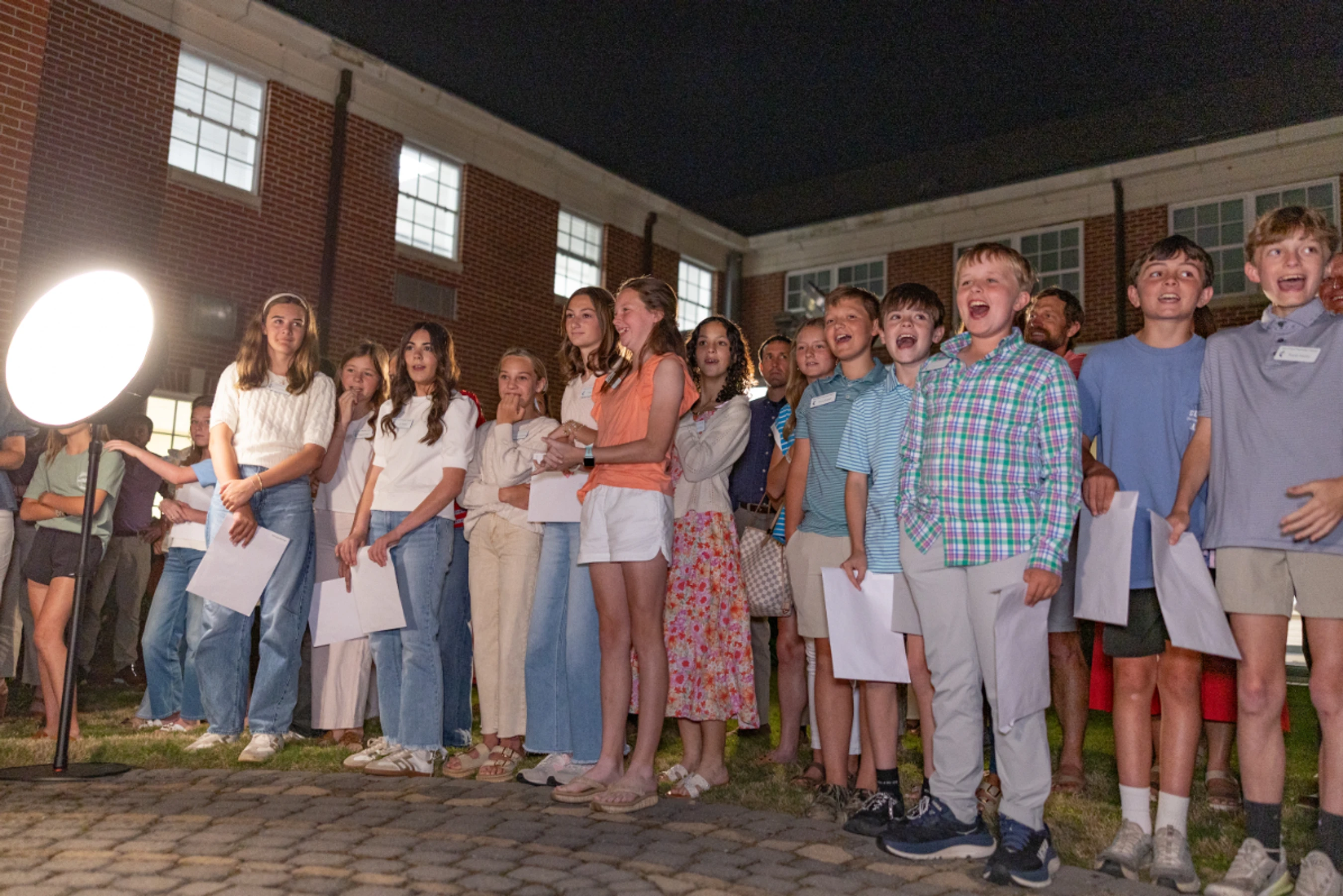 A large group of children and teenagers sing enthusiastically while standing outside a brick building at night, illuminated by a large circular light.