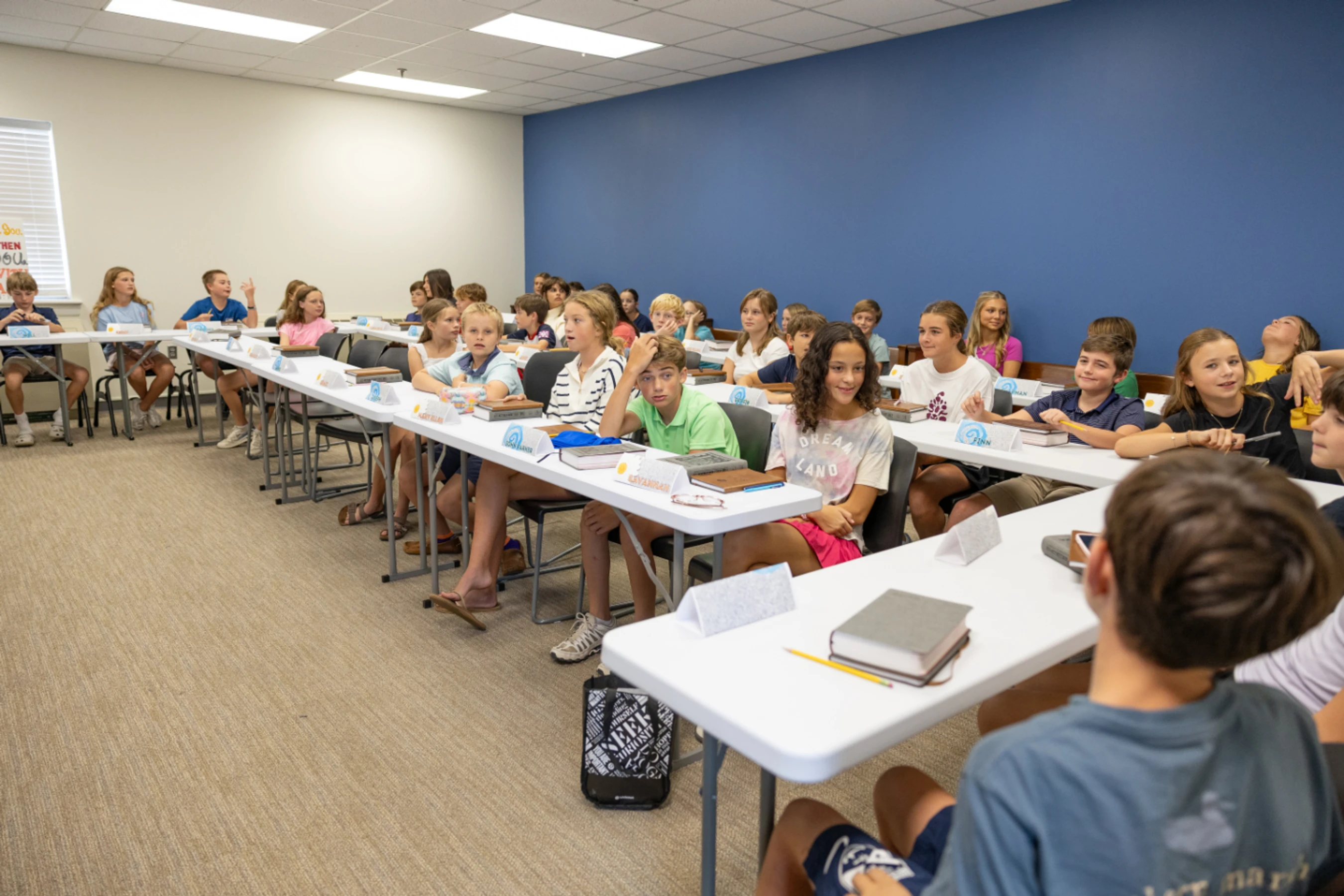 Many school children sit in two long rows of white tables in a classroom with a blue wall, looking towards the front of the room during a lesson.