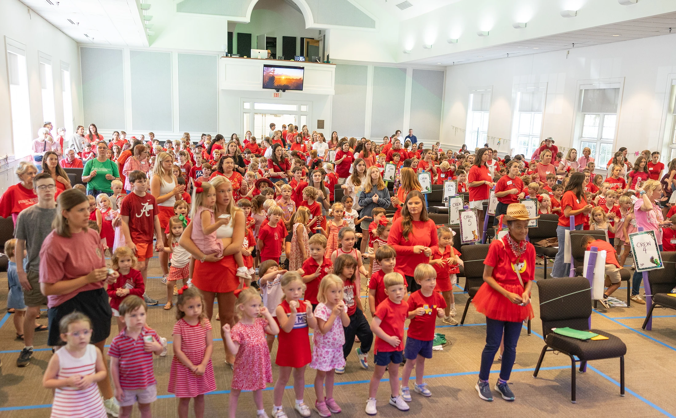 A large, bright hall filled with a crowd of children and adults, nearly all dressed in red, standing together for a community event.