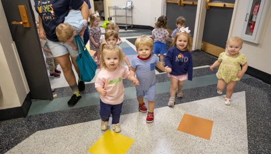 A group of young toddlers walking towards the camera down a school hallway. Some are holding hands and smiling, while an adult stands near a doorway.