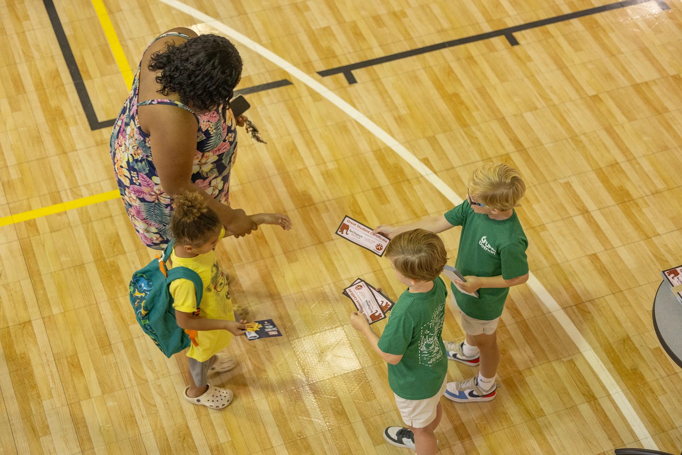 A high-angle photo shows a teacher handing certificates to three young children while standing on a polished wooden basketball court.