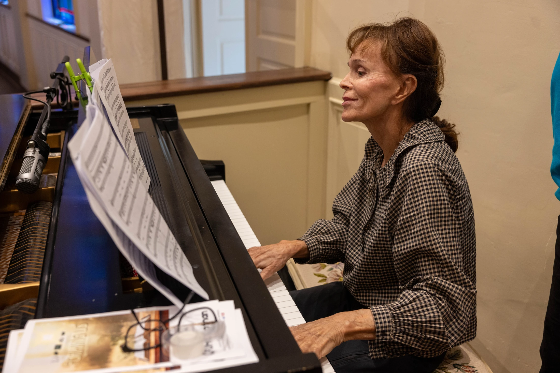 A side profile view of a woman with brown hair wearing a checkered shirt as she plays a black piano, looking at sheet music held up by clips.